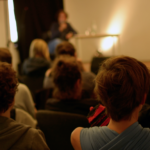 Menschen sitzen vor einer Bühne und hören einer Person zu.