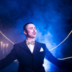 Mono steht vo einem Lichtkegel, trägt einen schwarzen Anzug mit weißem Hemd und goldener Fliege. Hinter ihm brennen 4 Kerzen und den Hintergrund durchziehen Nebelschwaden.